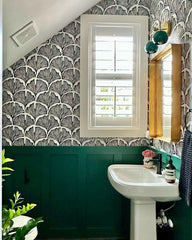 Colorful art deco inspired bathroom decor with graphic black and white patterned wallpaper, green wainscoting, and a two light green and brass wall sconce by Sazerac Stitches.