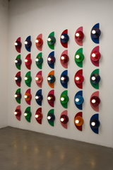 A grid of colorful Kin Sconces arranged in a gallery-style installation on a white wall. Each fixture features a unique trio of bold colors in a geometric, semi-circle design, casting a warm glow that highlights their sculptural forms.