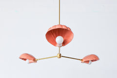 The Coquille Chandelier shown in a soft peach finish features three sculptural shell-shaped shades with white globe bulbs, mounted on a sleek brass three-arm structure. Suspended from a central brass rod, this ceiling fixture blends Art Deco elegance with whimsical, beach-inspired charm. The pastel tone and playful curves make it ideal for eclectic, coastal, or vintage-inspired spaces like dining rooms, nurseries, or boutique retail environments.