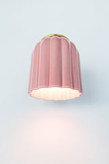 Close-up of the Marcella ceramic wall sconce in glazed pastel pink with a fluted shade and satin brass backplate, shown against a neutral background. This whimsical yet refined light fixture blends playful retro charm with elevated modern design, perfect for traditional, coastal, or cottagecore interiors seeking a soft pop of color and timeless silhouette.
