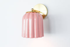 Close-up of the Marcella ceramic wall sconce in glazed pastel pink with a fluted shade and satin brass backplate, shown against a neutral background. This whimsical yet refined light fixture blends playful retro charm with elevated modern design, perfect for traditional, coastal, or cottagecore interiors seeking a soft pop of color and timeless silhouette.