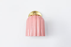 Close-up of the Marcella ceramic wall sconce in glazed pastel pink with a fluted shade and satin brass backplate, shown against a neutral background. This whimsical yet refined light fixture blends playful retro charm with elevated modern design, perfect for traditional, coastal, or cottagecore interiors seeking a soft pop of color and timeless silhouette.