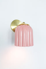 Close-up of the Marcella ceramic wall sconce in glazed pastel pink with a fluted shade and satin brass backplate, shown against a neutral background. This whimsical yet refined light fixture blends playful retro charm with elevated modern design, perfect for traditional, coastal, or cottagecore interiors seeking a soft pop of color and timeless silhouette.