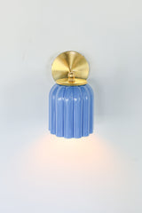 Marcella ceramic wall sconce in glazed sky blue with a fluted shade and brushed brass backplate, shown against a light gray background. This handcrafted fixture blends playful vintage style with modern sophistication, ideal for traditional, preppy, or coastal interiors seeking a pop of cheerful blue and timeless silhouette.