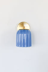 Marcella ceramic wall sconce in glazed sky blue with a fluted shade and brushed brass backplate, shown against a light gray background. This handcrafted fixture blends playful vintage style with modern sophistication, ideal for traditional, preppy, or coastal interiors seeking a pop of cheerful blue and timeless silhouette.