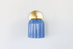 Marcella ceramic wall sconce in glazed sky blue with a fluted shade and brushed brass backplate, shown against a light gray background. This handcrafted fixture blends playful vintage style with modern sophistication, ideal for traditional, preppy, or coastal interiors seeking a pop of cheerful blue and timeless silhouette.