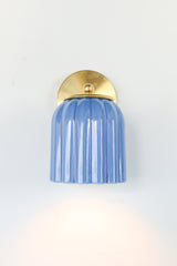 Marcella ceramic wall sconce in glazed sky blue with a fluted shade and brushed brass backplate, shown against a light gray background. This handcrafted fixture blends playful vintage style with modern sophistication, ideal for traditional, preppy, or coastal interiors seeking a pop of cheerful blue and timeless silhouette.