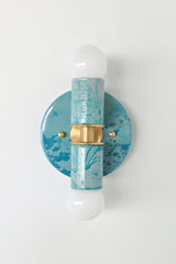 The Thalia Sconce in Teal and Grey Marbled finish is a bold, artistic wall light with two globe bulbs and a circular brass base. Featuring hand-applied marbled enamel in coastal-inspired hues, this sconce is perfect for adding a unique touch to bathroom lighting, powder rooms, or hall spaces.