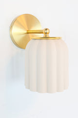 Marcella ceramic wall sconce in unglazed porcelain white with brushed brass hardware, showcasing a fluted, scalloped silhouette. This timeless fixture complements traditional, Southern-inspired, or coastal interiors with its soft matte finish and refined detailing—perfect for powder rooms, bathrooms, or hallways seeking classic charm with a sculptural edge.
