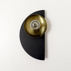 The Kin Sconce in matte black and polished brass features a bold semicircle backplate paired with a gleaming brass shade and socket. With its crisp geometric form and sculptural silhouette, this wall light is a nod to Bauhaus and Memphis design principles—clean lines, striking shapes, and elevated simplicity. The contrast of deep black and warm metallic makes it a standout accent in both minimal and maximalist interiors. Ideal for powder rooms, hallway vignettes, or flanking a bed in a modern, transitional,