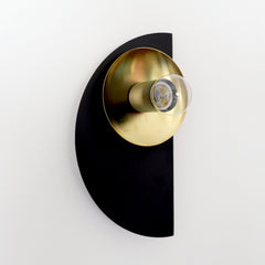 A graphic, architectural wall sconce from the Kin collection, shown in a dramatic black and brass colorway. The matte black semicircle backplate creates a bold silhouette against any wall, while the reflective brass shade adds a touch of glamour and warmth. Its Bauhaus-inspired geometry and sleek profile make it a versatile lighting choice—perfect for luxe bedrooms, artful entryways, or moody dining rooms with a sculptural edge. Designed to stand alone or in pairs, this fixture works beautifully in modern,