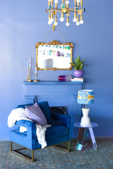 Blue Living room with Eclectic Decor details like an irridescent table, acrylic pill sculpture, antique mirror, brass evil eye chandelier, funky candle sticks, and antique fireplace grate.