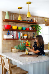 Modern kitchen remodel with brass pendant lights, neutral tile wall, wood floating shelves, and brightly colored accessories.