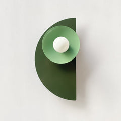 The Kin Sconce in a moody mix of olive green, vista green, and black, featuring layered geometric shapes and a round white bulb. A bold, sculptural wall fixture that blends Bauhaus formality with Memphis-inspired color blocking—perfect for interiors that lean modern, eclectic, retro, or maximalist. Works beautifully in creative offices, powder rooms, or as a statement-making hallway sconce.