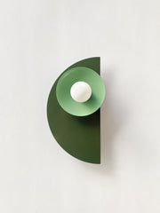 The Kin Sconce in a moody mix of olive green, vista green, and black, featuring layered geometric shapes and a round white bulb. A bold, sculptural wall fixture that blends Bauhaus formality with Memphis-inspired color blocking—perfect for interiors that lean modern, eclectic, retro, or maximalist. Works beautifully in creative offices, powder rooms, or as a statement-making hallway sconce.