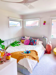 White bedroom with pops of color in musttard and bright pink.  Features a bright pink and brass wall sconce.  Used above a grey channel tufted day bed.