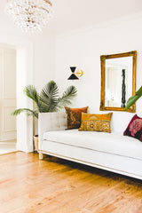 Pair of black Clancy sconces with warm brass accents frame a gilded mirror above a crisp white settee layered with jewel-toned velvet pillows. This living room combines midcentury modern lighting from Sazerac Stitches with global textiles and hardwood floors for a chic, collected, designer-approved look.