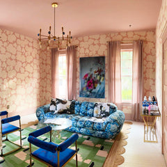 maximalist living room with pink floral walls, a brass evil eye chandelier, vintage floral patterned sofa, green geometric rug, and blue and brass sidechairs