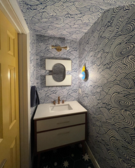 A peek into a whimsical bathroom reveals a Kin Sconce in citrus yellow and pastel blue tones, glowing against a bold navy-and-white wave wallpaper. Paired with celestial star floor tiles and a sculptural mirror, this space blends Memphis and Bauhaus influences with crisp, graphic precision.