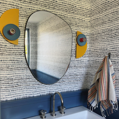 A Kin Sconce in yellow, periwinkle, and poolside blue adds a sculptural pop to this patterned bathroom, mounted beside a round mirror. The geometric fixture echoes the playfulness of the navy wave wallpaper and starry floor tile, proving how well it functions as a colorful, modern bathroom sconce.