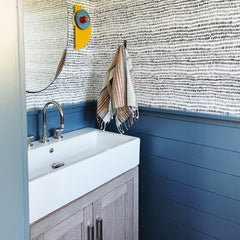 A Kin Sconce in yellow, periwinkle, and poolside blue adds a sculptural pop to this patterned bathroom, mounted beside a round mirror. The geometric fixture echoes the playfulness of the navy wave wallpaper and starry floor tile, proving how well it functions as a colorful, modern bathroom sconce.