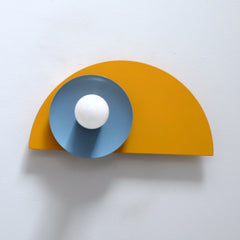 The Kin Sconce in mustard yellow, hydrangea blue, and cream, featuring bold Bauhaus-inspired geometry and playful Memphis design influences. This wall sconce showcases a striking half-circle backplate paired with a contrasting circular shade and round bulb, creating a sculptural, modern silhouette. A perfect statement light for geometric interiors, creative workspaces, or eclectic homes embracing color-forward design.