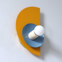 The Kin Sconce in mustard yellow, hydrangea blue, and cream, featuring bold Bauhaus-inspired geometry and playful Memphis design influences. This wall sconce showcases a striking half-circle backplate paired with a contrasting circular shade and round bulb, creating a sculptural, modern silhouette. A perfect statement light for geometric interiors, creative workspaces, or eclectic homes embracing color-forward design.