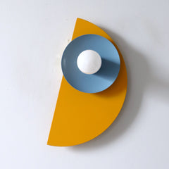 The Kin Sconce in a playful combination of mustard yellow, hydrangea blue, and cream, shown mounted on a white wall. Its bold half-circle backplate and angled disc shade create a sculptural, modern silhouette that blends Memphis-inspired geometry with mid-century modern color blocking—perfect for kids' rooms, creative studios, or eclectic interiors seeking a vibrant, artful lighting accent.