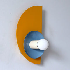 The Kin Sconce in mustard yellow, hydrangea blue, and cream, featuring bold Bauhaus-inspired geometry and playful Memphis design influences. This wall sconce showcases a striking half-circle backplate paired with a contrasting circular shade and round bulb, creating a sculptural, modern silhouette. A perfect statement light for geometric interiors, creative workspaces, or eclectic homes embracing color-forward design.