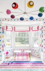 Bright kids’ playroom with rainbow Arcenciel Chandelier by Sazerac Stitches, colorful polka dot walls, hot pink roman shades, ghost chairs at a white kids’ table, rainbow striped rug, and built-in book displays. Bold, modern rainbow lighting for playrooms and nurseries.