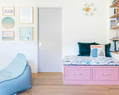 pastel living room with pink bench, natural wood shelving and framed art, and a white and brass loa carousel sconce by Sazerac Stitches