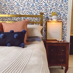 Modern bedroom design with a brass bed, white MCM loa sconce, blue pillow, blue and white traditional wallpaper, and an antique wood nightstand.