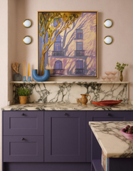 Kitchen vignette with four Sazerac Stitches Abeille wall lights in brass with white globe diffusers flanking a large framed painting on soft pink walls; deep purple shaker cabinets and island, veined marble countertop and backsplash with a shallow display ledge; styled with a blue sculptural vase, tapered candles, coupe glasses, herbs, and a red tray; warm ambient glow, classic meets modern palette, handmade customizable lighting from New Orleans.