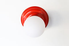 Poppy Orange Red Retro Style  flushmount ceiling light or wall sconce with a frosted white globe.  Stepping back into a bygone era with a touch of modern minimalism, the Abeille Sconce or Flushmount is an artistic blend of form and function. This wall sconce doubles as a flushmount ceiling light, making it a versatile lighting solution for any space. Designed akin to a beehive, its charismatic shape is complemented by curated color options that add a playful element to its retro appeal. 