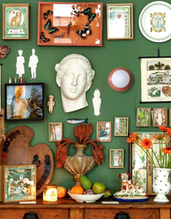 Eclectic gallery wall on deep green paint with salon-style arrangement of vintage frames, butterfly specimens, botanical charts, plaster bust, and small curios above a rustic wood sideboard; bowl of fruit, lit candle, and orange gerbera daisies in a dotted vase add warmth; centered on the wall is a Sazerac Stitches Abeille wall light in peach with a white globe diffuser, a compact low-profile accent that suits maximalist, collected interiors; handmade, customizable color.