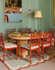 Adele Chandelier in polished brass with flame orange scalloped bobeches, styled above a round wood dining table with red bamboo dining chairs, green painted walls, and colorful modern artwork. This scalloped chandelier adds retro charm, sculptural drama, and a pop of color to dining rooms, blending traditional and modern lighting design. Perfect for eclectic, colorful interiors that need a statement chandelier.