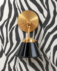 A black wall sconce with a glossy cone shade and raw brass hardware. A swivel knuckle with a wing nut on the round backplate lets you tilt the shade. It hangs on black and white zebra stripe wallpaper with a soft woven texture. Use it as a vanity light in a powder room, a bedside reading light, or a pair flanking a bar. Black, white, and warm brass make a crisp high contrast mix that suits Hollywood Regency, maximalist, and glam powder room ideas.