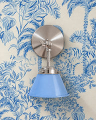 A light blue wall sconce with a glossy cone shade and polished nickel hardware. A swivel knuckle with a wing nut on the round backplate lets you tilt the shade. It hangs on cream wallpaper painted in blue palms, leafy jungle foliage, and hidden tigers. Use it as a bedside sconce, a vanity light by a bathroom mirror, or a reading light in a nursery. Blue on blue keeps it fresh and calm, a good fit for coastal, chinoiserie, and blue and white bedroom or bathroom decor.