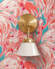 A cream wall sconce with a glossy cone shade and raw brass hardware. A swivel knuckle with a wing nut on the round backplate lets you tilt the shade. It sits on loud flamingo wallpaper in coral, hot pink, and turquoise with sweeping painterly feathers. The quiet cream shade lets the print do the talking. Hang it by a bathroom mirror as a vanity light or beside a bed. It suits tropical, Palm Beach, and maximalist powder room ideas, and very New Orleans ones too.