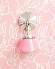 A bubblegum pink wall sconce with a glossy cone shade and polished nickel hardware. A swivel knuckle with a wing nut on the round backplate lets you tilt the shade. It hangs on pink wallpaper covered in loose white sketched bunnies, pink on pink with a bright silver backplate. Use it as a bedside reading light in a girls room or nursery, or a vanity light in a pink powder room. Sweet but not fussy, it suits playful kids room decor, pink nursery ideas, and whimsical modern rooms.