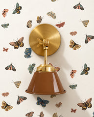 A caramel brown wall sconce with a glossy cone shade and raw brass hardware. A swivel knuckle with a wing nut on the round backplate lets you tilt the shade. It sits on cream wallpaper scattered with illustrated moths and butterflies in dusty pink, teal, and gold. Hang it beside a bed, over a desk, or next to a bathroom mirror as a vanity light. Warm brown and brass pick up the earthy tones in the print, so it suits vintage, cottage, and cozy eclectic bedrooms and reading nooks.