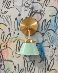A mint green wall sconce with a glossy cone shade and raw brass hardware. A swivel knuckle with a wing nut on the round backplate lets you tilt the shade. Here it hangs on painterly bunny wallpaper with black brushstroke rabbits over washes of blue, pink, and yellow. Use it as a bedside reading light in a nursery or kids room, or a vanity light in a powder room. Mint and brass keep a busy pattern light and playful, a good fit for whimsical, grandmillennial, and colorful kids room decor.
