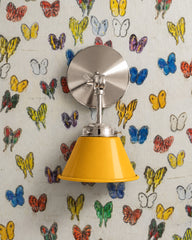 A golden yellow wall sconce with a glossy cone shade and polished nickel hardware. A swivel knuckle with a wing nut on the round backplate lets you tilt the shade. It hangs on cream wallpaper covered in painterly butterflies in yellow, cobalt, red, purple, and green. Use it as a bedside reading light, a desk lamp in a kids room, or a vanity light. Sunny yellow pulls from the print and keeps it cheerful, a good fit for colorful kids rooms, eclectic bedrooms, and art filled maximalist decor.