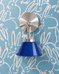 A cobalt blue wall sconce with a glossy cone shade and polished nickel hardware. A swivel knuckle with a wing nut on the round backplate lets you tilt the shade. It sits on light blue wallpaper covered in loose white sketched bunnies. Deep cobalt against soft blue makes a fresh tonal mix. Use it as a bedside reading light in a boys room or nursery, or a vanity light in a kids bathroom. It suits blue nursery ideas, playful kids room decor, and whimsical coastal rooms.