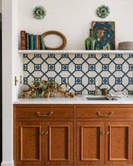 Traditional kitchen or butler’s pantry with warm wood cabinets, marble countertops, brass hardware, and a blue and cream geometric tile backsplash. A pair of Aged Green Octavia sconces add a small, sculptural pop of color above the floating marble shelf, styled with vintage books, art, glassware, candlesticks, and collected objects. This feels classic, grandmillennial, and slightly English country, perfect for customers looking for decorative kitchen wall sconces, vintage-inspired lighting, blue and white t