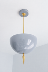 Handmade grey pendant light that reads as a soft neutral with a sculptural edge — a high-gloss powder-coated dome in warm dove grey, lifted by a slim brass stem and a hand-turned brass finial. The color is calm and adaptable: it reads gentle greige in daylight and softens under lamplight, so it settles into any palette while the glossy finish and brass keep it from feeling flat. A quiet fixture that goes with everything — white walls, wood, color, or a moody backdrop. Perfect over a kitchen island, dining n
