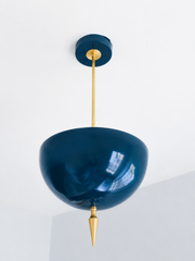 Handmade lagoon pendant light in a rich jewel-toned blue-green — a high-gloss powder-coated dome poised between teal and navy, grounded by a slim brass stem and a hand-turned brass finial. The saturated color reads slate-blue in daylight and deepens to an inky peacock tone under lamplight, so it works like a jewel-toned neutral against warm wood, white walls, or patterned wallpaper. Perfect over a kitchen island, powder room, or entryway that needs a hit of depth. Shop handmade blue pendant lighting, glossy