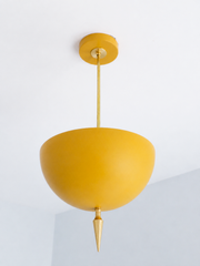 Handmade mustard pendant light with a soft matte finish that makes yellow feel warm and lived-in rather than bright — a powder-coated dome in deep ochre-gold, anchored by a slim brass stem and a hand-turned brass finial that drops to a fine point. The flat, chalky surface reads earthy and vintage: it leans golden in daylight and turns honey-warm under lamplight, so it settles easily into a room with wood, wicker, or cream tones as well as a bolder color-drenched space. Perfect over a kitchen island, a break
