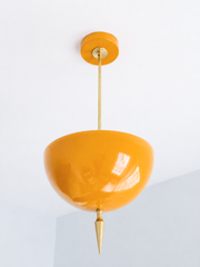 Alexander Pendant in glossy marigold-orange powder-coat, shown at a three-quarter angle with light pooling across the domed metal shade, slim brass stem, and turned brass finial spike below.
