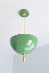 Handmade green pendant light in a soft, glossy vista green — a high-gloss powder-coated dome in a muted sage-celadon tone, lifted by a slim brass stem and a hand-turned brass finial. The color is calm and organic: it reads dusty sage in daylight and warms under lamplight, so it settles easily against brass, natural wood, cream, or a botanical wallpaper without ever feeling loud. Perfect over a kitchen island, a breakfast nook, a sunroom, or an entryway that could use a grounded touch of green. Shop handmade
